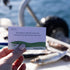 Hand holds a Getting Lost Fishing Edition card choosing who selects bait for the next cast on a boat, Adventure Awaits