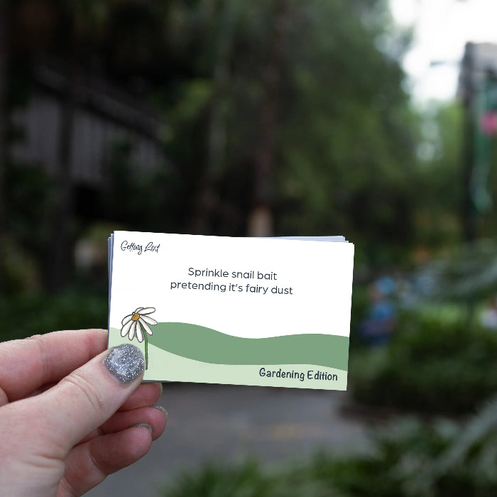 Hand holds a Getting Lost Gardening Edition card prompting snail bait sprinkled like fairy dust, Adventure Awaits