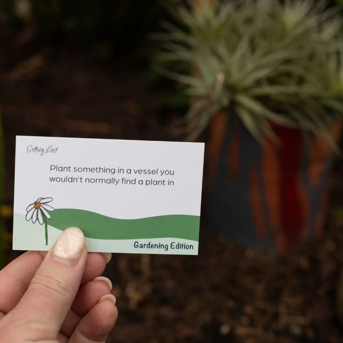 Hand holds a Getting Lost Gardening Edition card prompting planting in an unusual vessel, Adventure Awaits