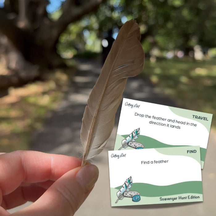 Hand holding a feather beside Scavenger Hunt Edition cards on a shady park path — Adventure Awaits