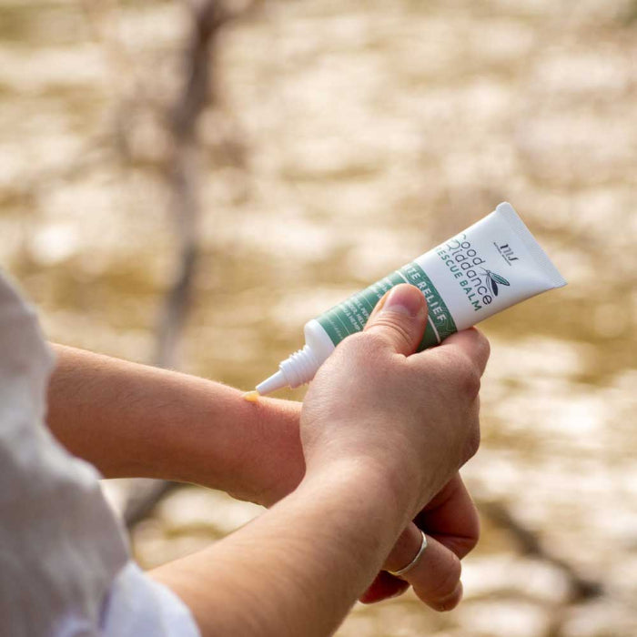 Hand applying a tube of Good Riddance Rescue balm to a person's arm with a blurred natural background