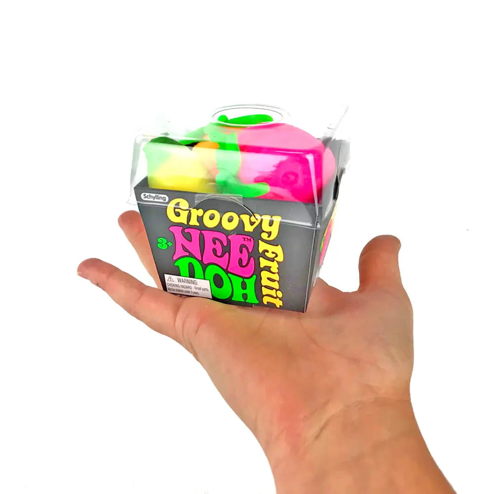 A hand holding a packaged novelty product labeled 'Groovy Nee Doh Fruit' against a plain white background. The product inside the clear plastic box appears to be a bright pink and green squishy toy or novelty item.