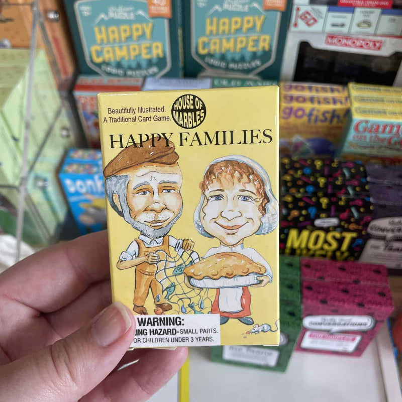 Hand holding a 'Happy Families' card game box with 'Happy Camper' and other games in the background.