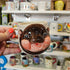 Person holding a mug with a baby pygmy hippo design in a store with various mugs on display.