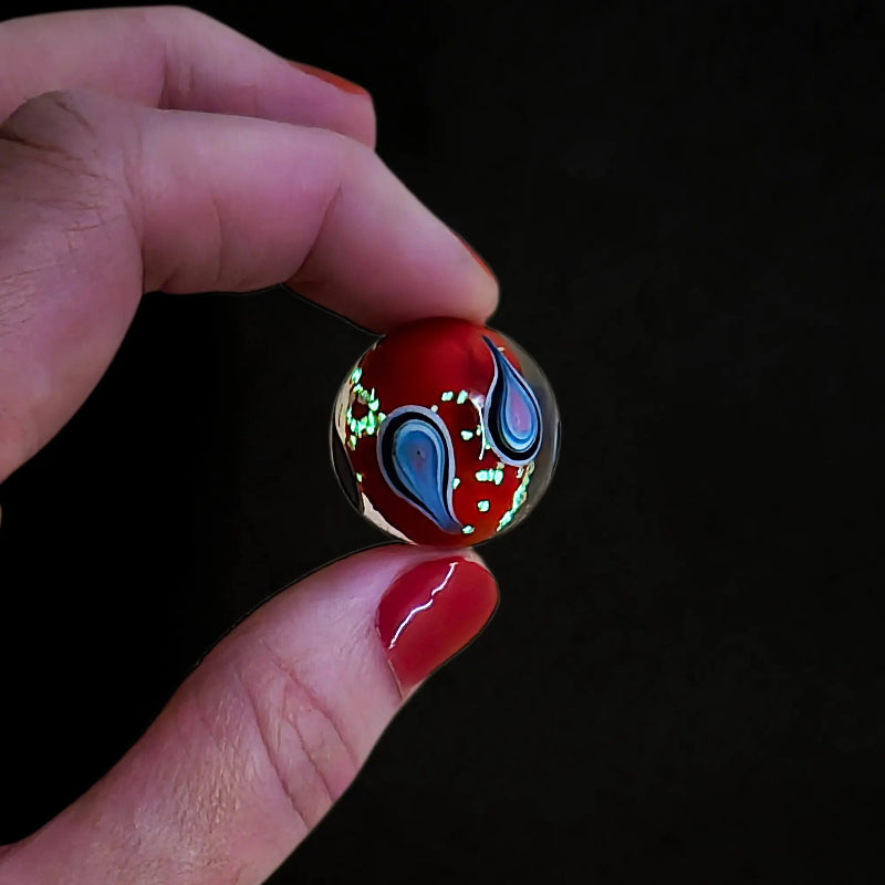 Hand holding a red glow-in-the-dark glass marble with blue and pink teardrop swirls, Adventure Awaits