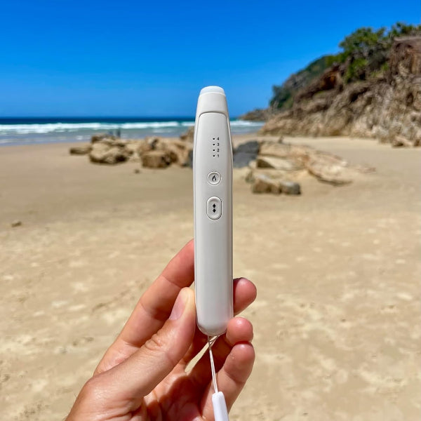 Hand holding a white electronic insect relief pen device on a beach with ocean and cliffs in the background