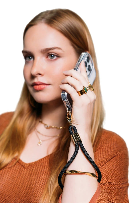 Woman takes a call with her phone secured by a black wrist strap with gold clasp, Adventure Awaits
