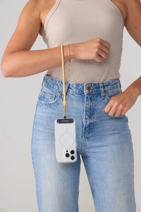 Woman wears a tan phone wrist strap carrying her phone with gold clasp beside blue jeans, Adventure Awaits