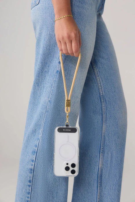 Woman carries a phone by a tan wrist strap with gold clasp beside blue jeans, Adventure Awaits