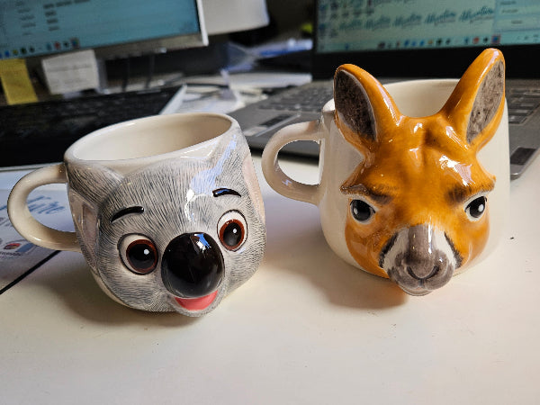 Two animal-themed mugs, one a koala and the other a kangaroo, on a desk.