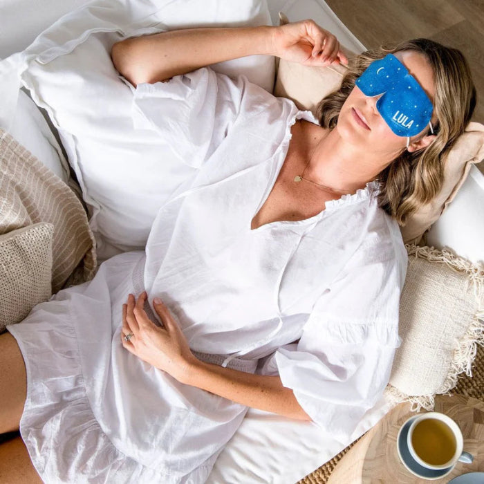 Woman relaxing on a couch wearing Lula self warming eye mask, designed to soothe tired eyes and promote restful sleep and relaxation at home or travel Adventure Awaits