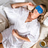 Woman relaxing on a couch wearing Lula self warming eye mask, designed to soothe tired eyes and promote restful sleep and relaxation at home or travel Adventure Awaits