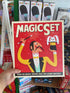 Hand holding a 'Magic Set' book with a colorful cover featuring a magician, surrounded by more books in the background.