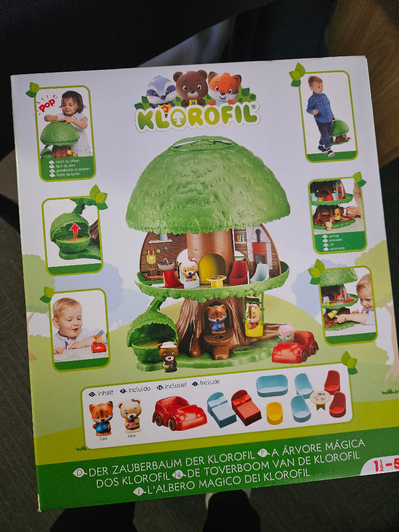 The retail packaging box for the Klorofil Magic Tree toy set, photographed on a dark surface. The box features a colorful illustration of the green treehouse with a brown trunk, surrounded by small inset photos of children playing with the toy and a row of included pieces at the bottom.
