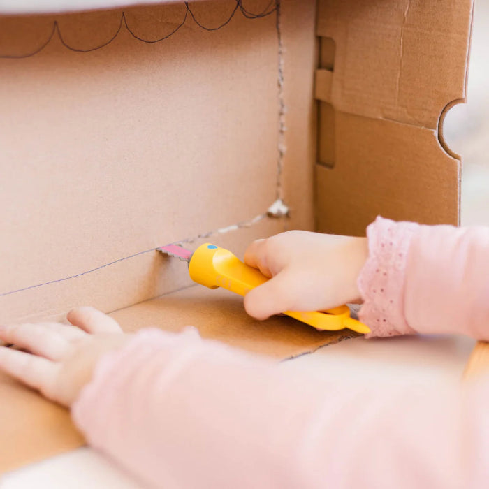 Child uses a yellow Makedo Safe-Saw to cut cardboard horizontally along a drawn line during creative play — Adventure Awaits