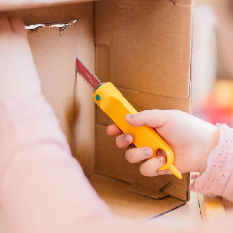 Child uses a yellow Makedo Safe-Saw to cut cardboard along a drawn line during creative play — Adventure Awaits
