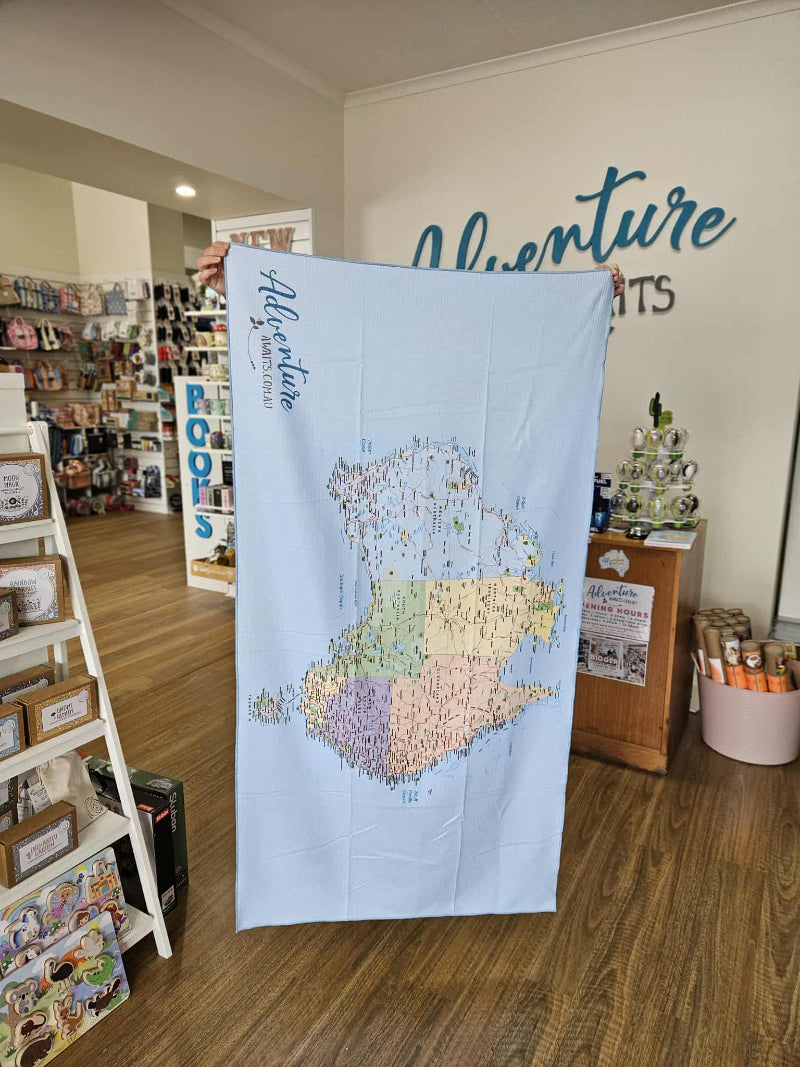 A vertically held version of the map towel showing Australia printed upside down, displaying the Adventure Awaits logo at the top. The towel is held by someone hidden behind it in a retail setting.