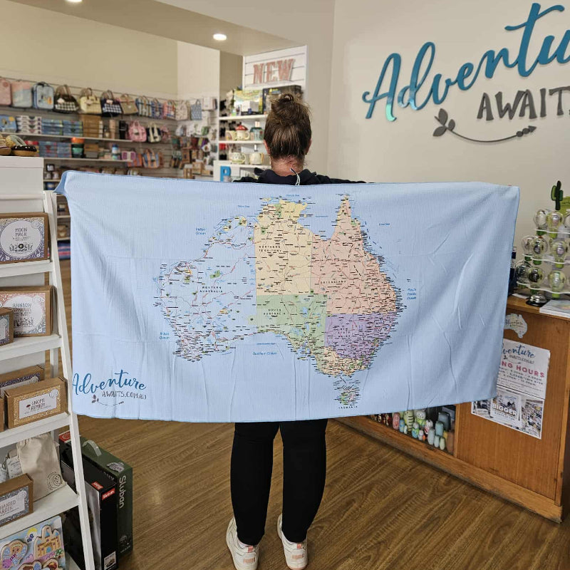 A person standing indoors holding out a large light blue towel featuring a full map of Australia. The towel reads "Adventure Awaits" in the corner and is displayed inside a retail shop.
