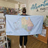 A person standing indoors holding out a large light blue towel featuring a full map of Australia. The towel reads "Adventure Awaits" in the corner and is displayed inside a retail shop.