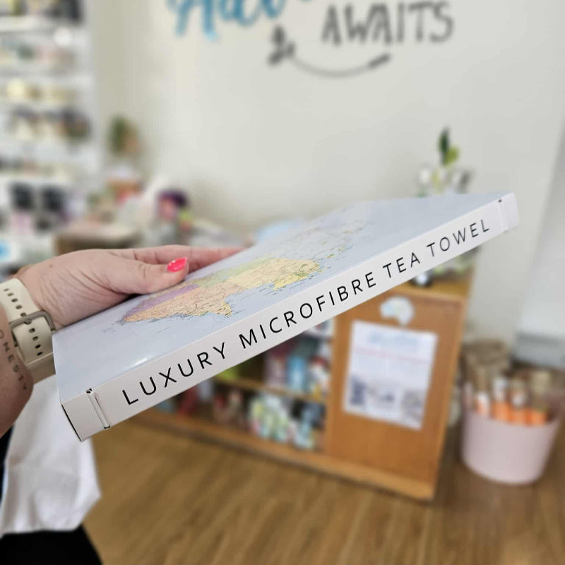 Person holding a luxury microfibre tea towel packaging with 'Adventure Awaits' in the background.