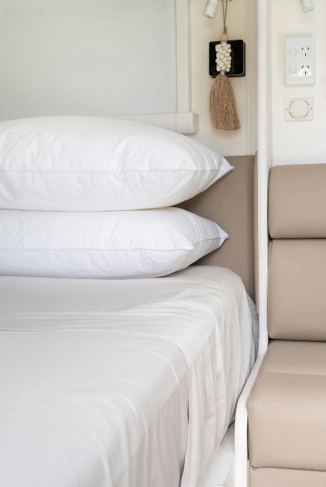 Two stacked white pillows on neatly made bed beside caravan wall, clean and comfortable bedding setup for travel Adventure Awaits