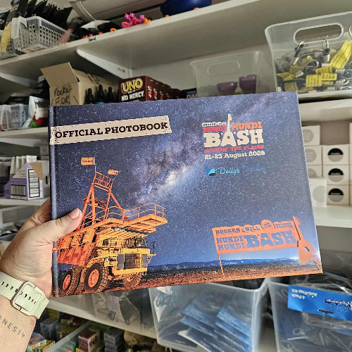 Hand holding an 'Official Photobook' for 'Bash' event against a cluttered shelf background