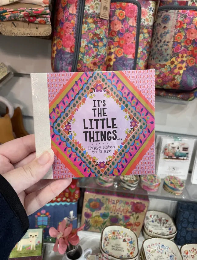 Hand holding It’s the Little Things happy notes booklet in colourful gift shop display — Adventure Awaits