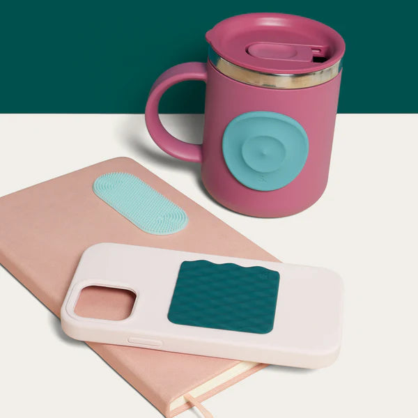 Pink mug with teal lid on a white surface, next to a phone case with teal silicone fidget sticker by speks