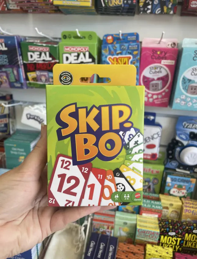Hand holding a Skip-Bo card game box in front of a colourful games display — Adventure Awaits