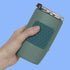 A hand holding a matte sage green insulated tumbler with a dark teal textured grip and a stainless steel lid, set against a solid light blue background.