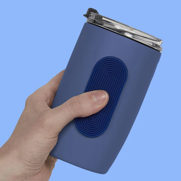 A hand holding a blue insulated tumbler with a dark blue textured oval silicone fidget sticker by speks on the front and a clear lid, set against a solid light blue background.