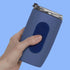 A hand holding a blue insulated tumbler with a dark blue textured oval silicone fidget sticker by speks on the front and a clear lid, set against a solid light blue background.
