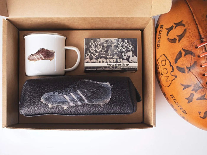 The Footballers Gift set with enamel mug, footballers soap and black boot-print wash bag beside a vintage football — Adventure Awaits