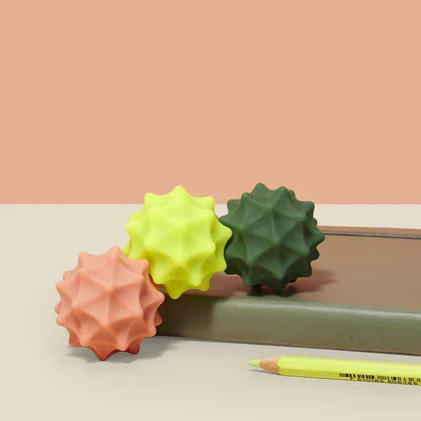 Three textured stress balls in pink, green, and yellow on a light surface with a pen.