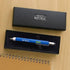 A blue and silver mechanical pencil with a ruler scale design and 'Men's Republic' branding, displayed inside its black presentation box on a light wood tabletop next to a spiral notebook.