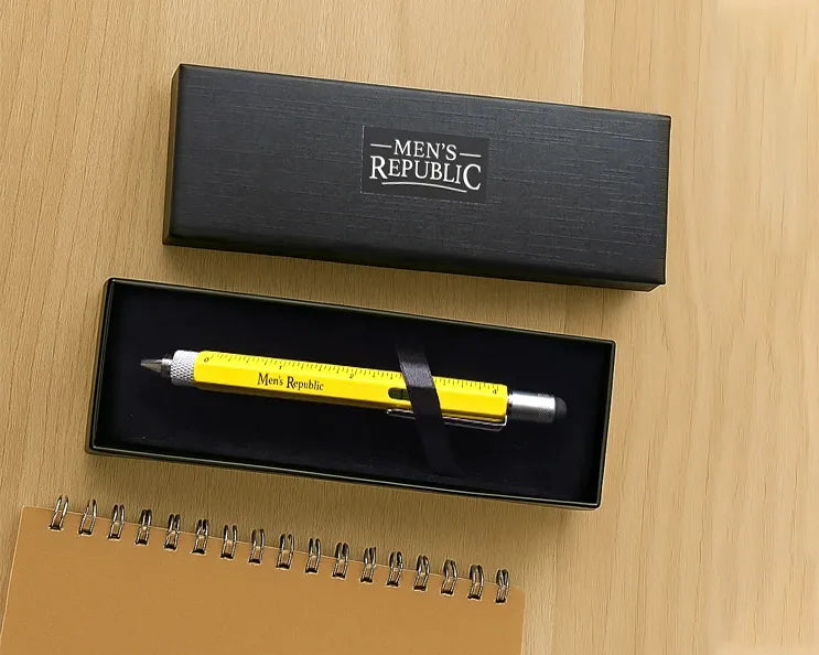 A yellow and silver Men's Republic branded retractable pen with a measuring scale design, presented in an open black gift box on a light wood tabletop.