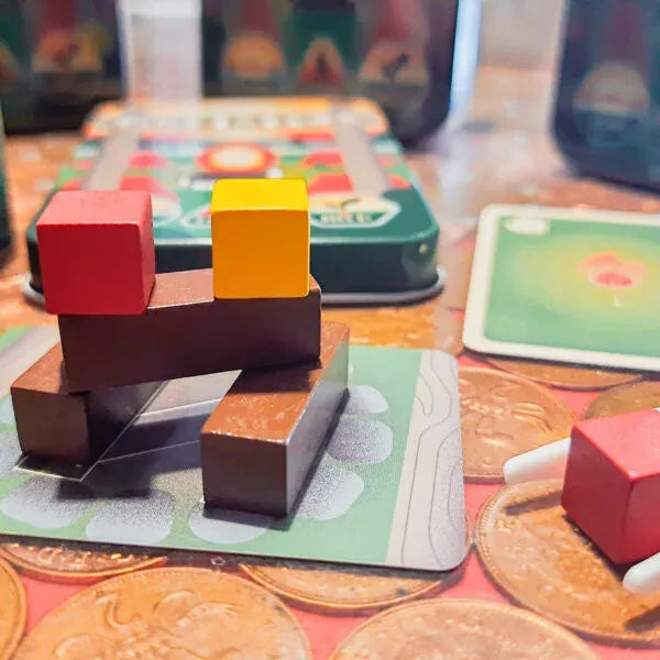 A close-up of a wooden block stacking toy or game setup on a tabletop, with red and yellow wooden blocks on dark brown supports, surrounded by cards, coins, and a green tin in the background.
