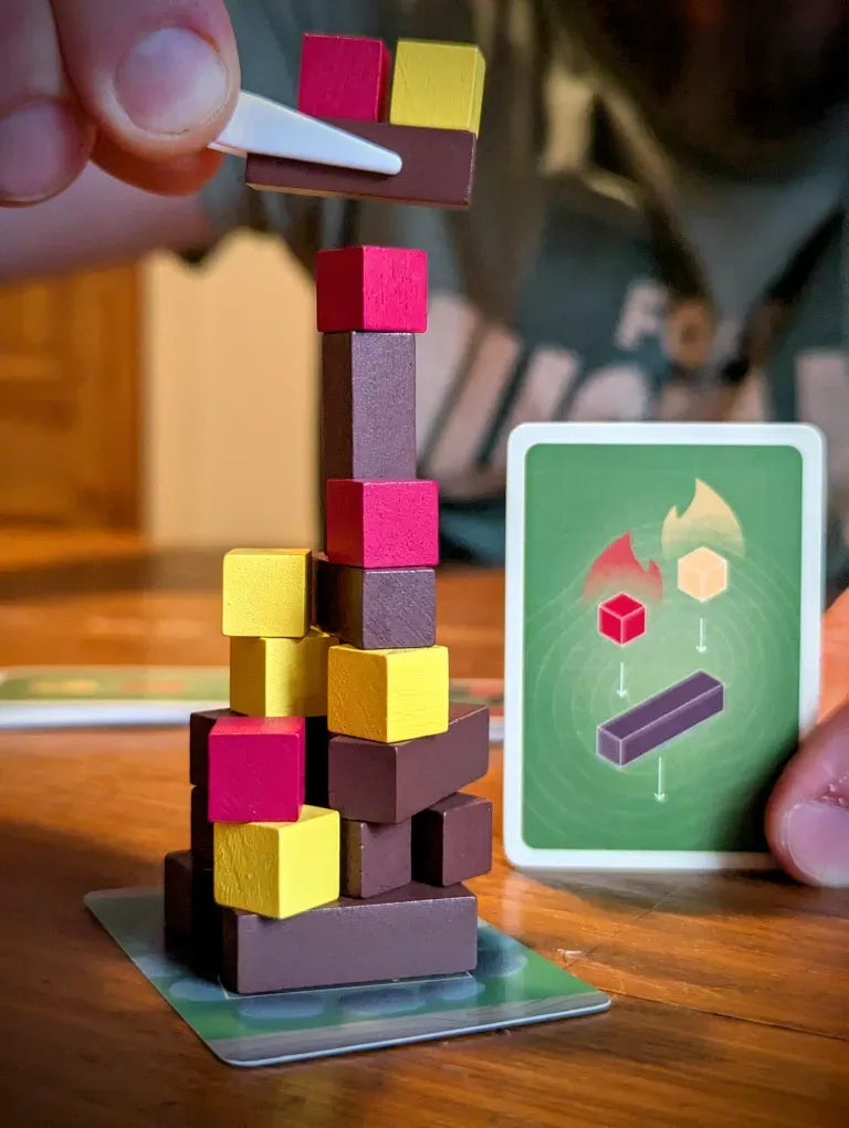 A close-up of a hand placing a wooden block onto a tall, colorful tower game, with a green card held in the other hand on a wooden table.