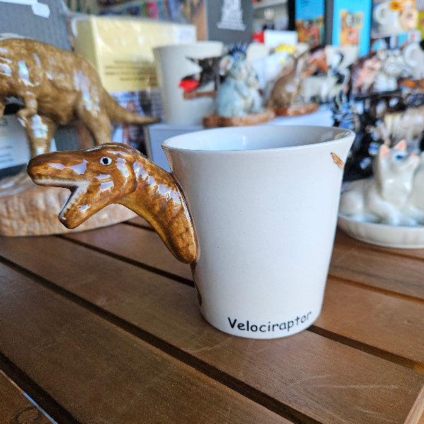 White mug with 'Velociraptor' printed on it attached to a wooden surface with a dinosaur figurine below.