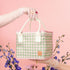 Green gingham handbag held by a gloved hand with flowers on a pink background
