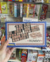 Hand holding a Philos Rummy tile game box in front of a colourful games display — Adventure Awaits