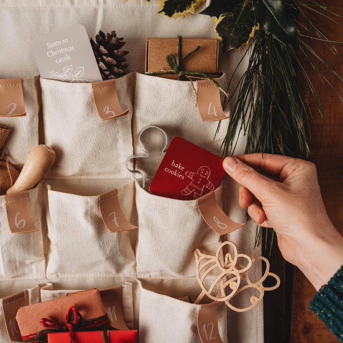 Hand placing a red bake cookies Mindful Advent Activity Card into a fabric advent calendar — Adventure Awaits