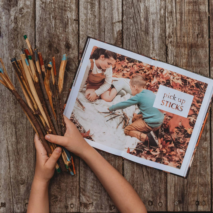 Child holding painted sticks beside an open Wild Child nature craft book on a rustic table — Adventure Awaits