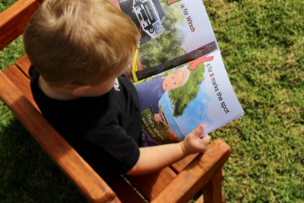 young child sitting outdoors reading ABC for Little Offroaders book, exploring letters and adventure scenes while seated on wooden chair in grass Adventure Awaits