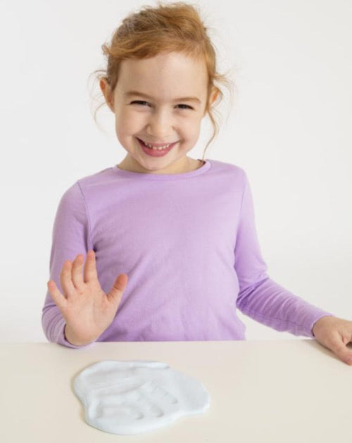 girl in purple shirt sitting behind handprint impression made on white clay 