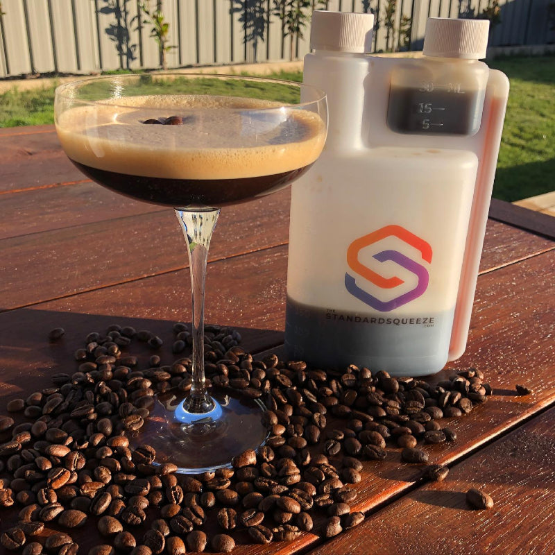Espresso martini beside Standard Squeeze Bottle on outdoor table scattered with coffee beans — Adventure Awaits