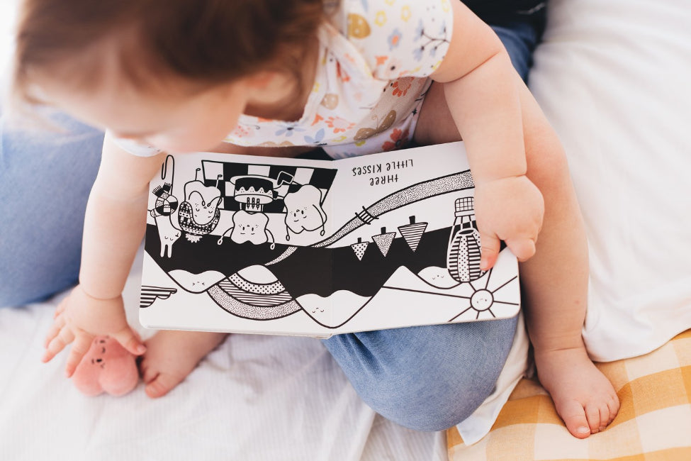 Baby looking at black and white Ten Little Kisses For You board book on bed — Adventure Awaits