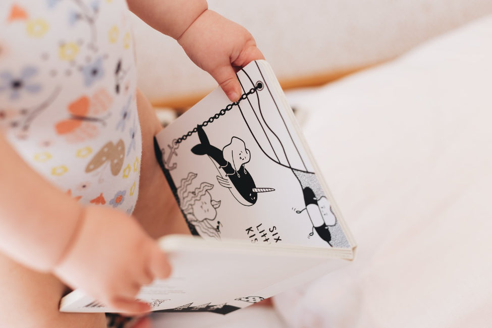 Baby holding open Ten Little Kisses For You board book on soft bedding — Adventure Awaits