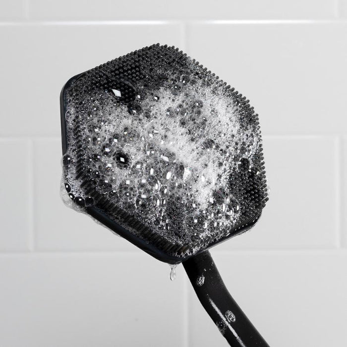 close p of front of tooletries back scrubber covered in suds held up against white shower